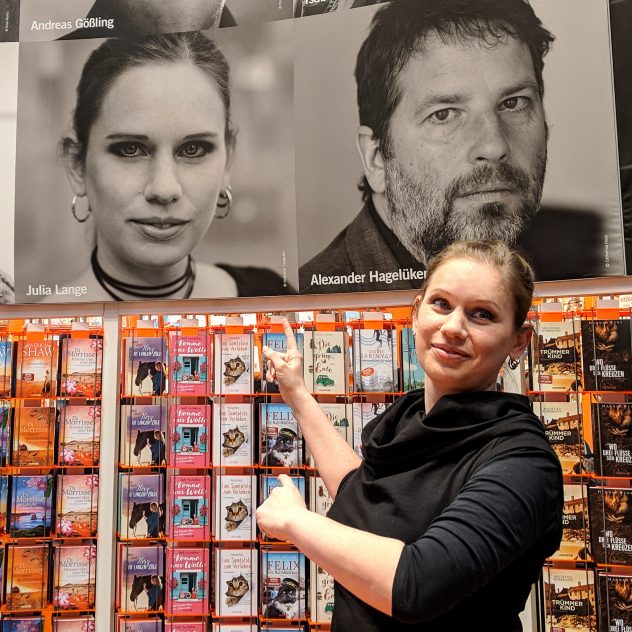 Julia Lange vor einer Bilderwand am Knaur-Stand, wo ihr Foto groß neben anderen Autorenkollegen.